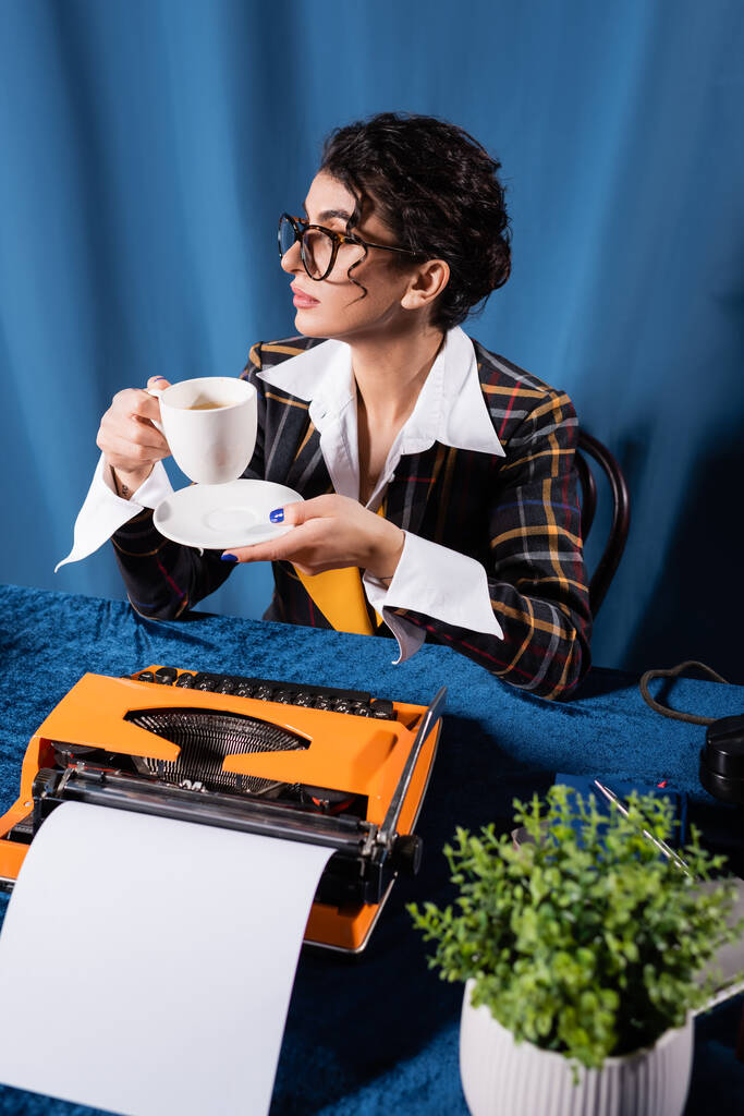 Moda gazetecisi elinde kahve bardağıyla mavi arka planda klasik daktilonun yanında oturuyor. - Fotoğraf, Görsel