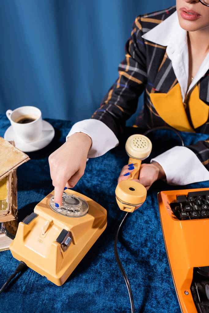 μερική άποψη της κομψής γυναίκας καλώντας στο τηλέφωνο κοντά στη γραφομηχανή και φλιτζάνι καφέ σε μπλε φόντο - Φωτογραφία, εικόνα