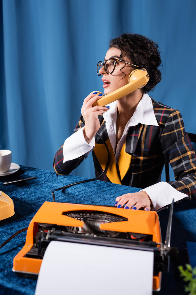 journaliste surpris parlant au téléphone près de la machine à écrire sur fond bleu avec draperie - Photo, image