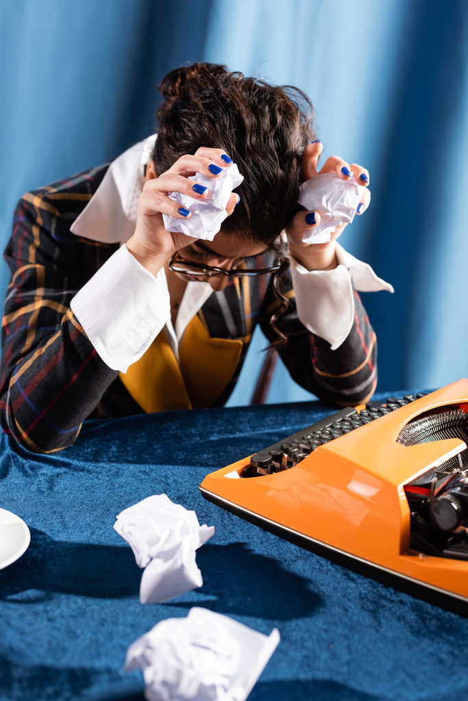 depresivní novinář sedí se skloněnou hlavou a zmačkaný papír v blízkosti vintage psacího stroje na modrém pozadí - Fotografie, Obrázek