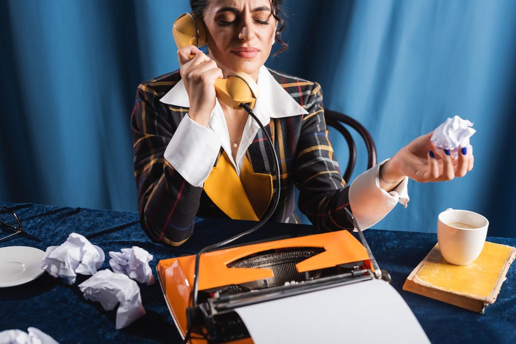 mulher descontente com os olhos fechados segurando papel amassado e falando ao telefone no fundo azul - Foto, Imagem