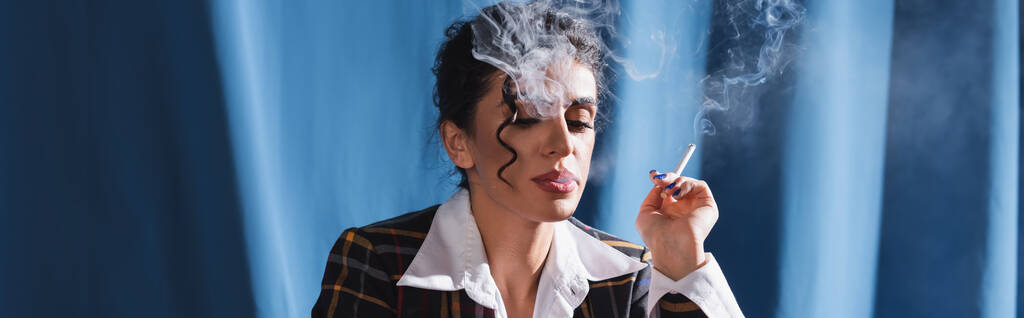 Esmer, eski elbiseli bir kadın mavi arka planda sigara içiyor. - Fotoğraf, Görsel