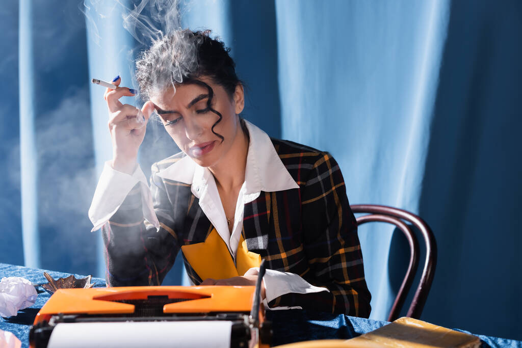 ヴィンテージタイプライターでタイプし青を背景に喫煙するスタイリッシュな女性 - 写真・画像