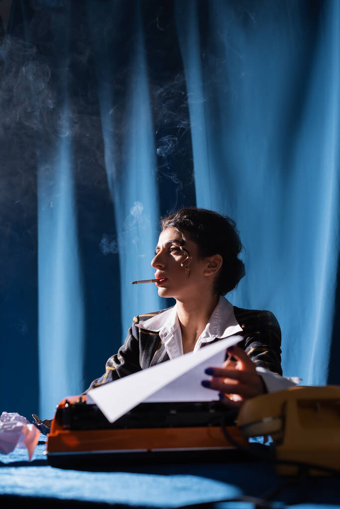 stijlvolle vrouw houden papier in de buurt wazig typemachine terwijl roken op de achtergrond met blauwe draperieën - Foto, afbeelding