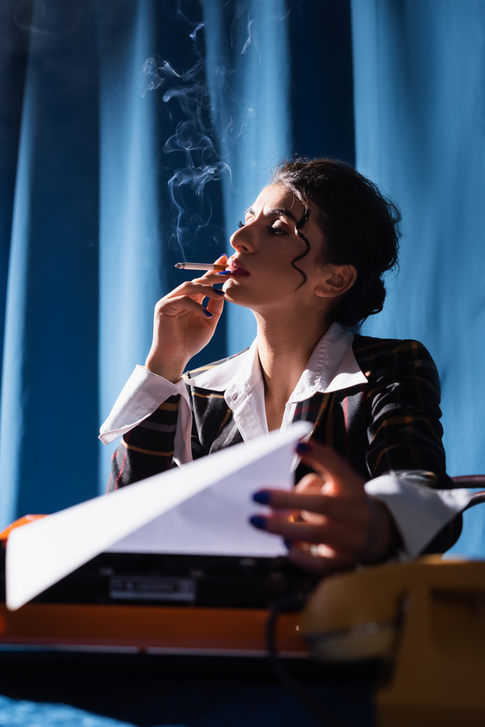 стильная женщина в стиле ретро курит рядом с пишущей машинкой на синем фоне с занавеской - Фото, изображение