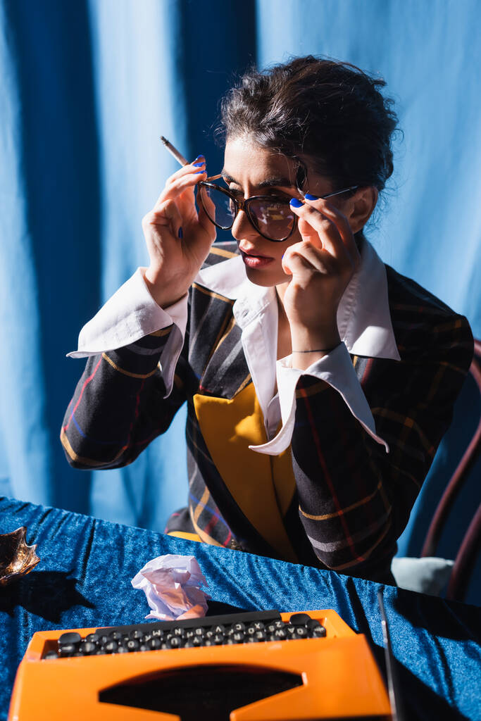 retro styl žena s cigaretou nastavení brýle v blízkosti psacího stroje na modrém pozadí - Fotografie, Obrázek