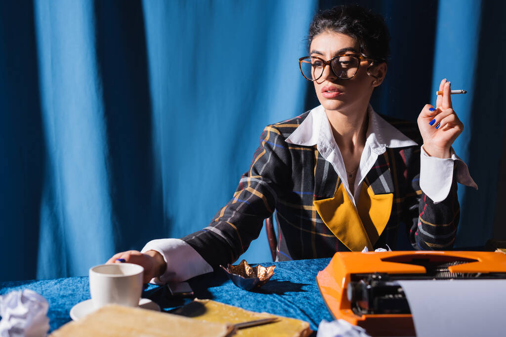 Журналистка винтажного стиля с сигаретой рядом с чашкой кофе и пишущей машинкой на синем фоне - Фото, изображение