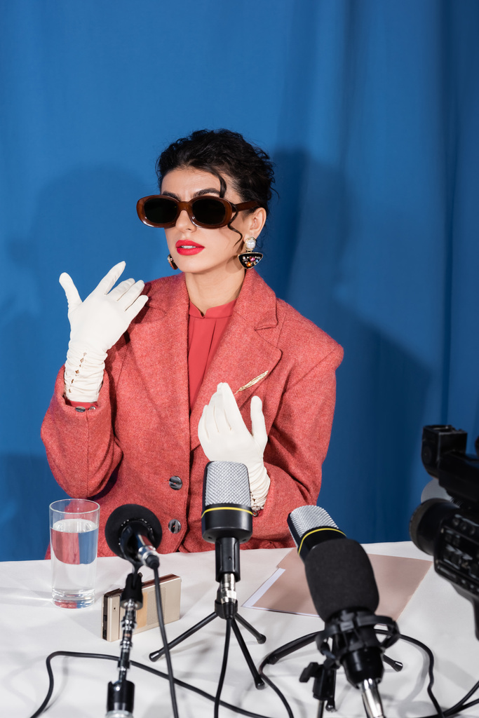 stylish woman in vintage sunglasses and white gloves gesturing during interview on blue background - Photo, Image