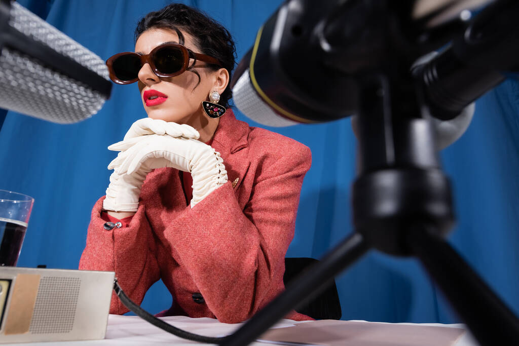 розмиті мікрофони біля стильної жінки в старовинних сонцезахисних окулярах та білих рукавичках на синьому фоні
 - Фото, зображення
