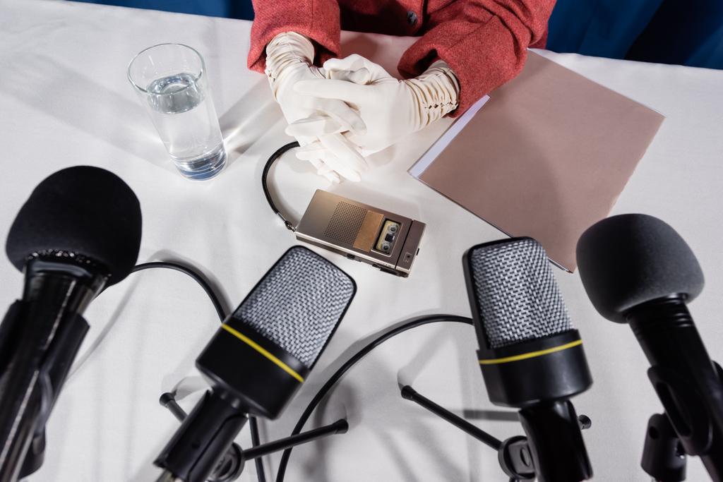 μερική άποψη της γυναίκας σε λευκά γάντια, μικρόφωνα και συσκευή εγγραφής φωνής σε λευκό τραπέζι   - Φωτογραφία, εικόνα