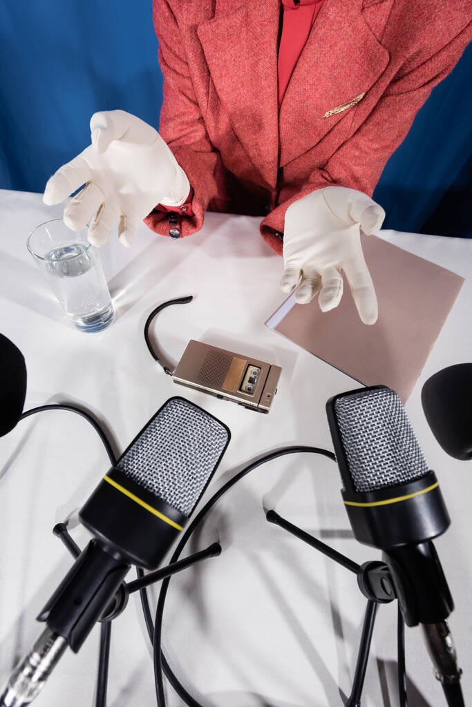 Mikrofonların üst görüntüsü, ses kaydedici ve mavi arka planda beyaz eldivenli bir kadının yanında bir bardak su. - Fotoğraf, Görsel