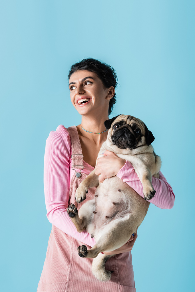 sonriente mujer mirando hacia otro lado y sosteniendo perro pug aislado en azul - Foto, imagen