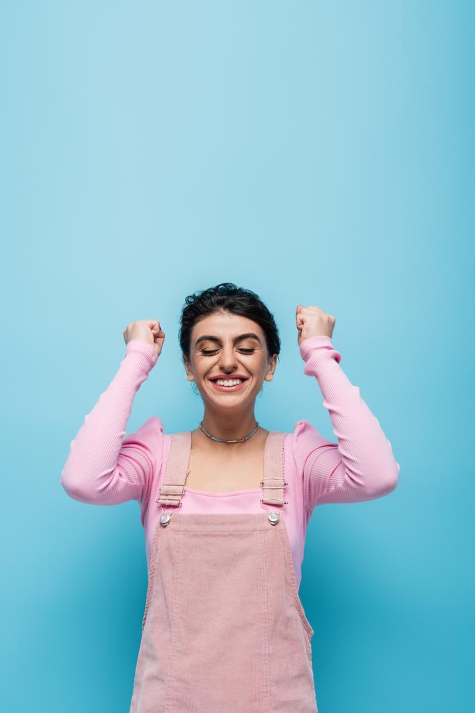 excited woman with closed eyes showing win gesture isolated on blue - Photo, Image