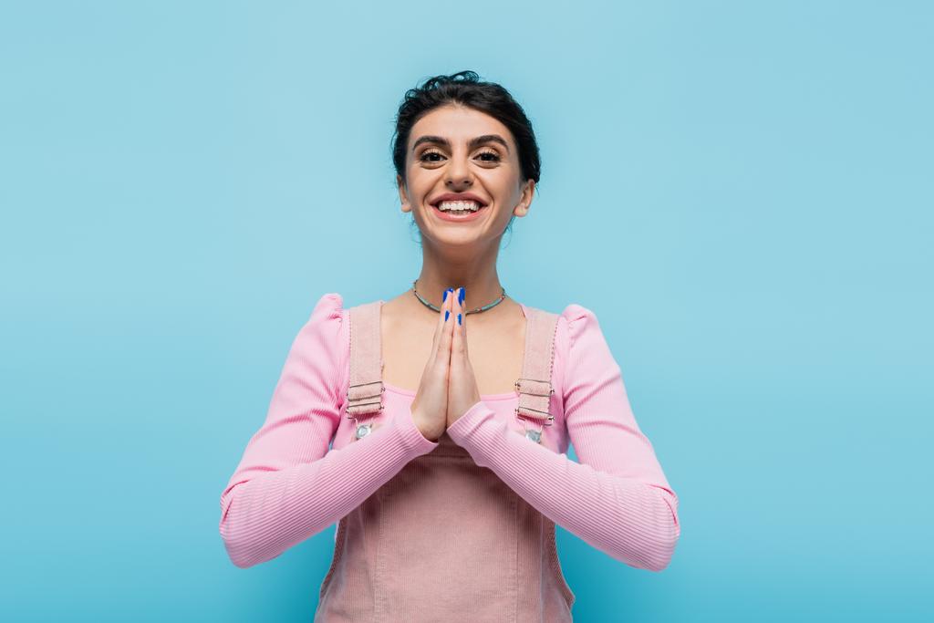 young brunette woman showing gratitude gesture and smiling at camera isolated on blue - Photo, Image