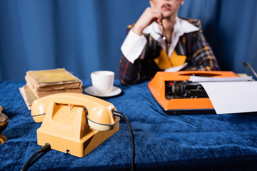 visão parcial da jornalista borrada perto do telefone vintage, máquina de escrever e livros sobre toalha de mesa de veludo azul - Foto, Imagem