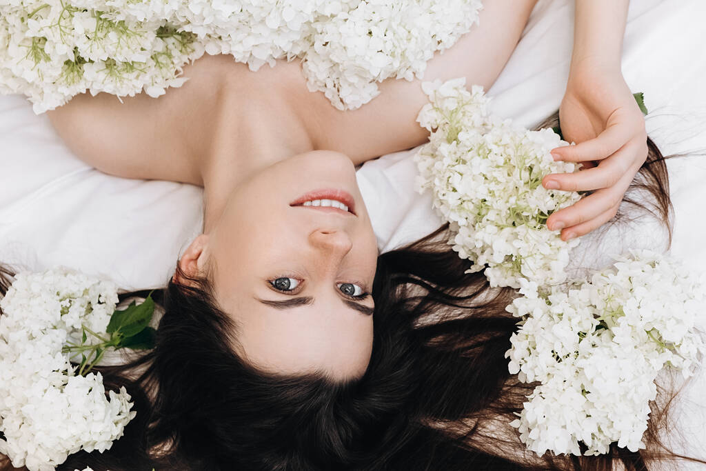 Ξέγνοιαστο πρωινό. Κοντινό πλάνο πορτρέτο των χαρούμενων νεαρή χαμογελαστή γυναίκα 20 ετών χαλαρώνοντας σε ζεστό λευκό κρεβάτι στο σπίτι, θετική κοιτάζοντας κάμερα, απολαμβάνει φρέσκα λουλούδια άνοιξη. Έννοια συναισθημάτων - Φωτογραφία, εικόνα