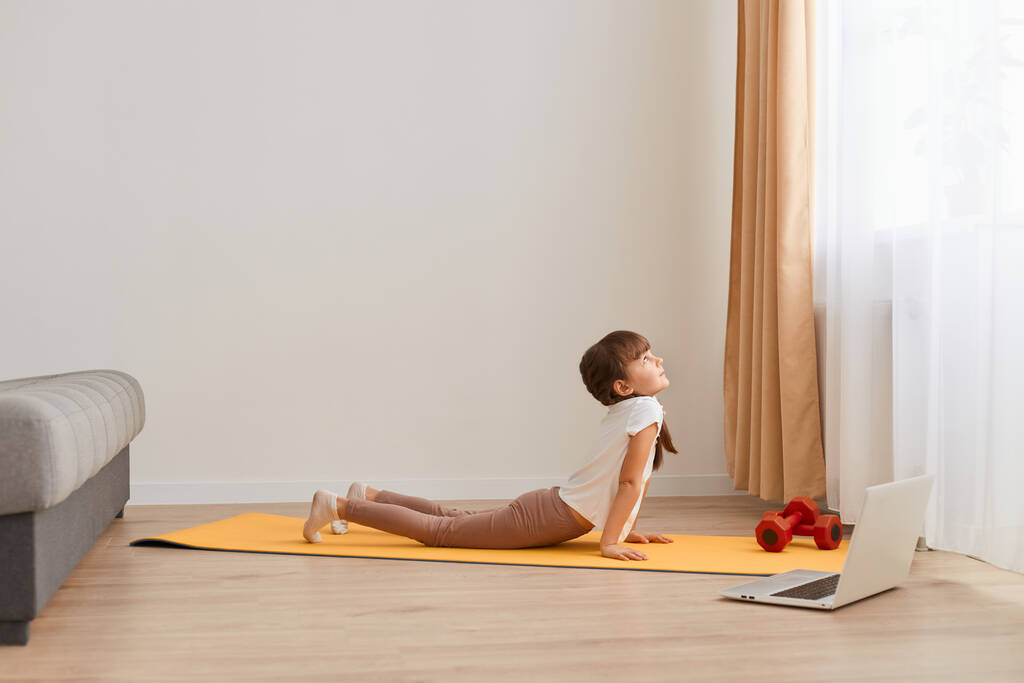 Indoor shot of little cute girl practicando yoga pose on a mat indoor, estirando su cuerpo, usando ropa casual, teniendo trenzas, posando en la sala de estar de luz cerca de la ventana. - Foto, Imagen