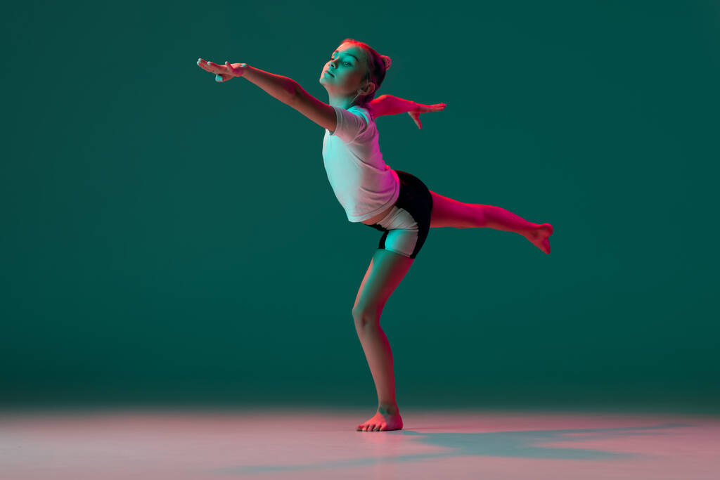 Bailando. Niña flexible, entrenamiento de artista de gimnasia rítmica aislado en fondo de estudio verde en luz rosa neón. Gracia en movimiento, acción. Hacer ejercicios de flexibilidad. - Foto, imagen