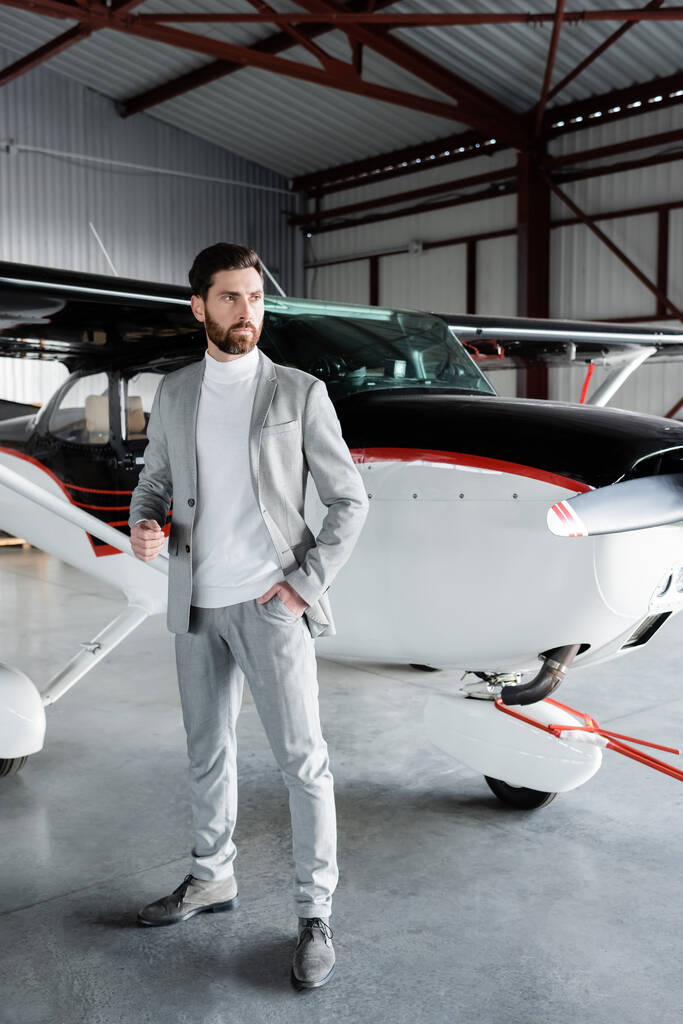 полная длина уверенного бизнесмена в костюме, стоящего с рукой в кармане рядом с современным вертолетом - Фото, изображение