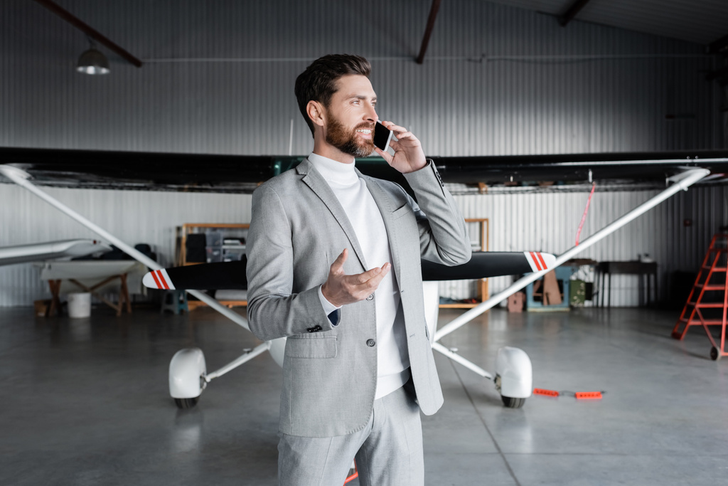 hombre feliz en ropa formal hablando en el teléfono inteligente y el gesto cerca de helicóptero moderno - Foto, imagen
