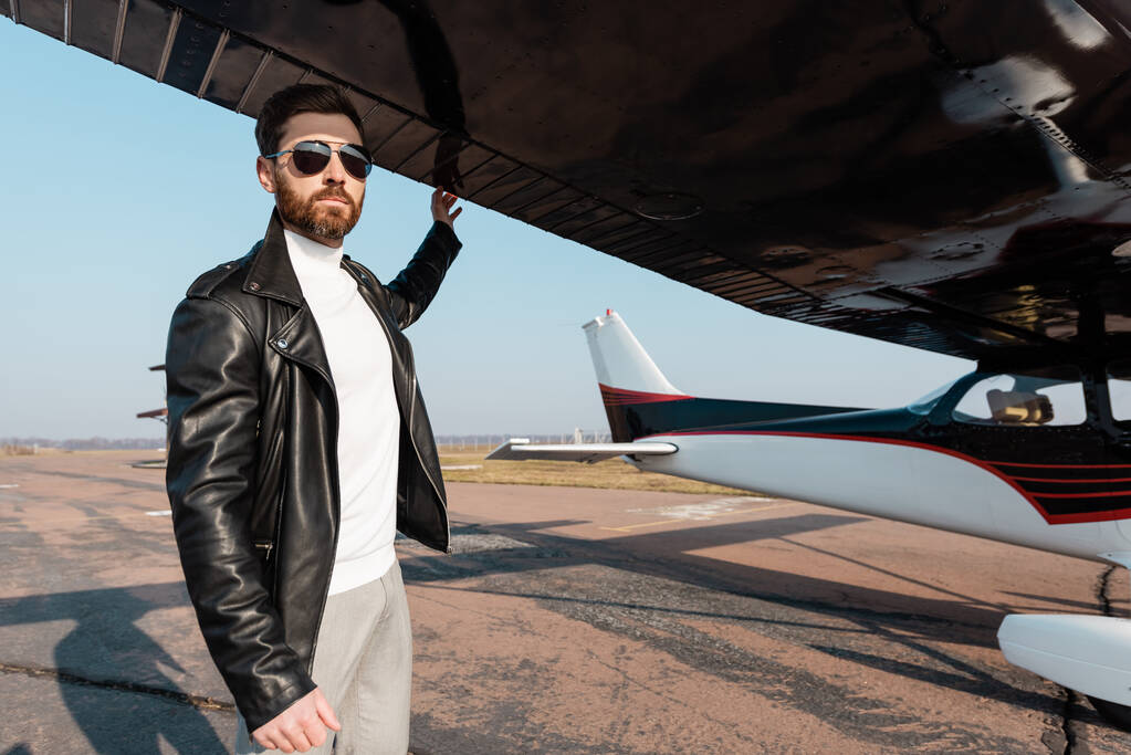 Güneş gözlüklü ve deri ceketli sakallı pilot uçak kanadının altında duruyor. - Fotoğraf, Görsel