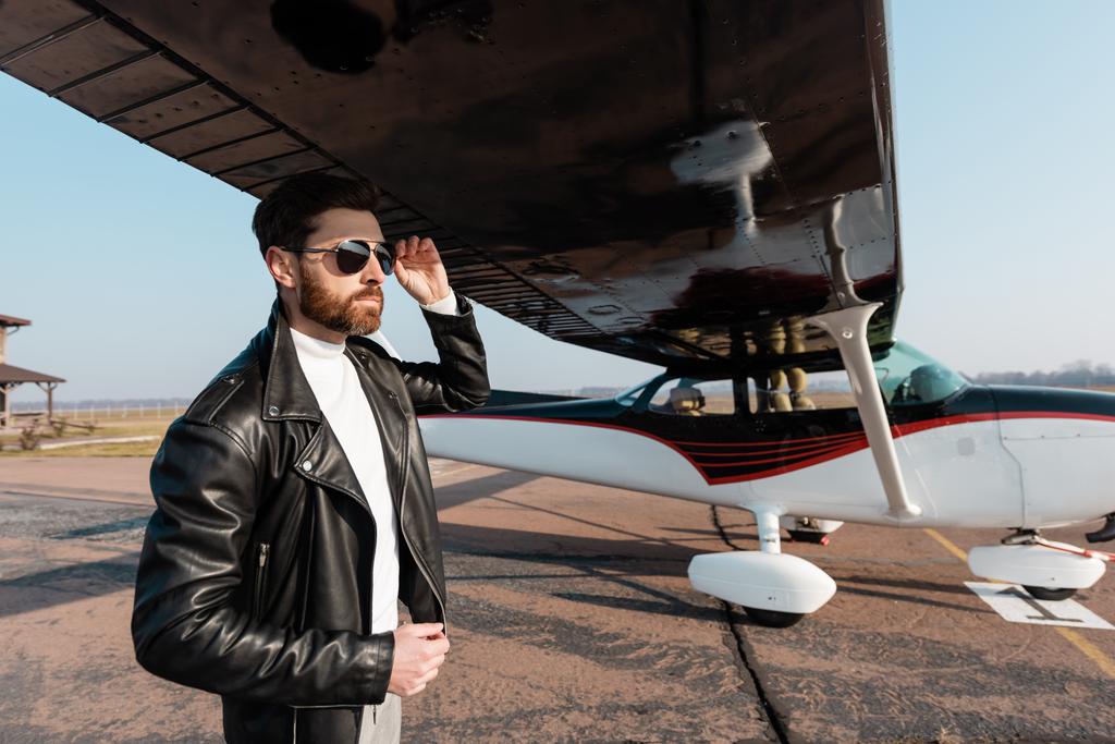 pilota barbuto in giacca di pelle regolazione occhiali da sole sotto ala aereo - Foto, immagini
