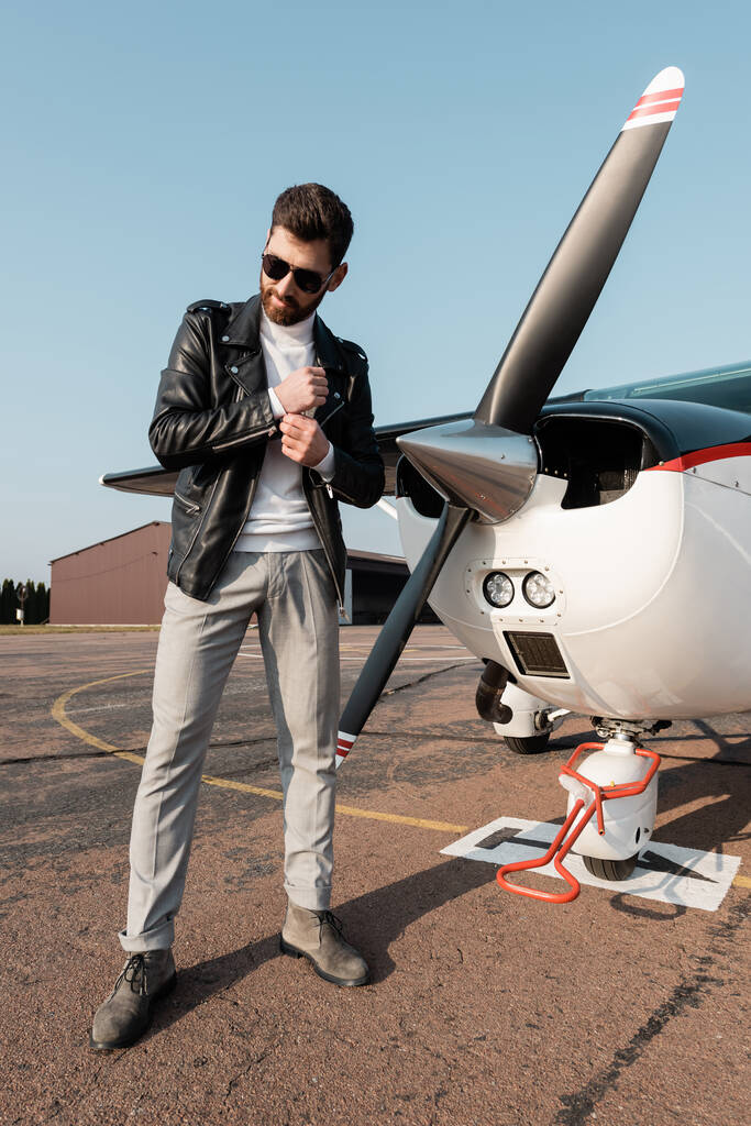 полная длина стильного пилота в кожаной куртке и солнцезащитных очках улыбаясь, стоя рядом с самолетом  - Фото, изображение