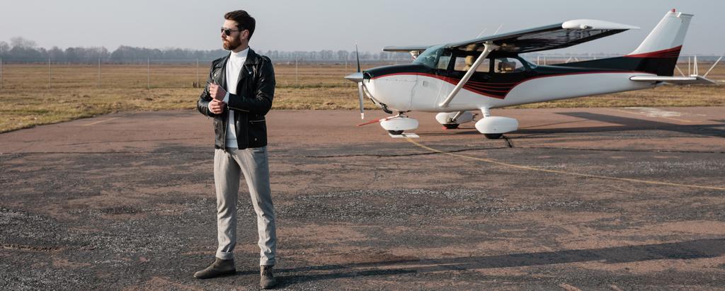 полная длина стильного пилота в кожаной куртке и солнцезащитных очках, стоящих возле вертолета, баннер - Фото, изображение