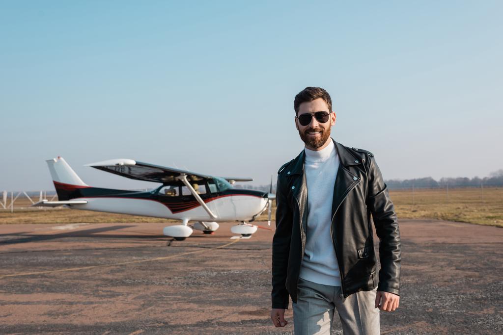 happy pilot in stylish leather jacket and sunglasses smiling near blurred helicopter - Photo, Image