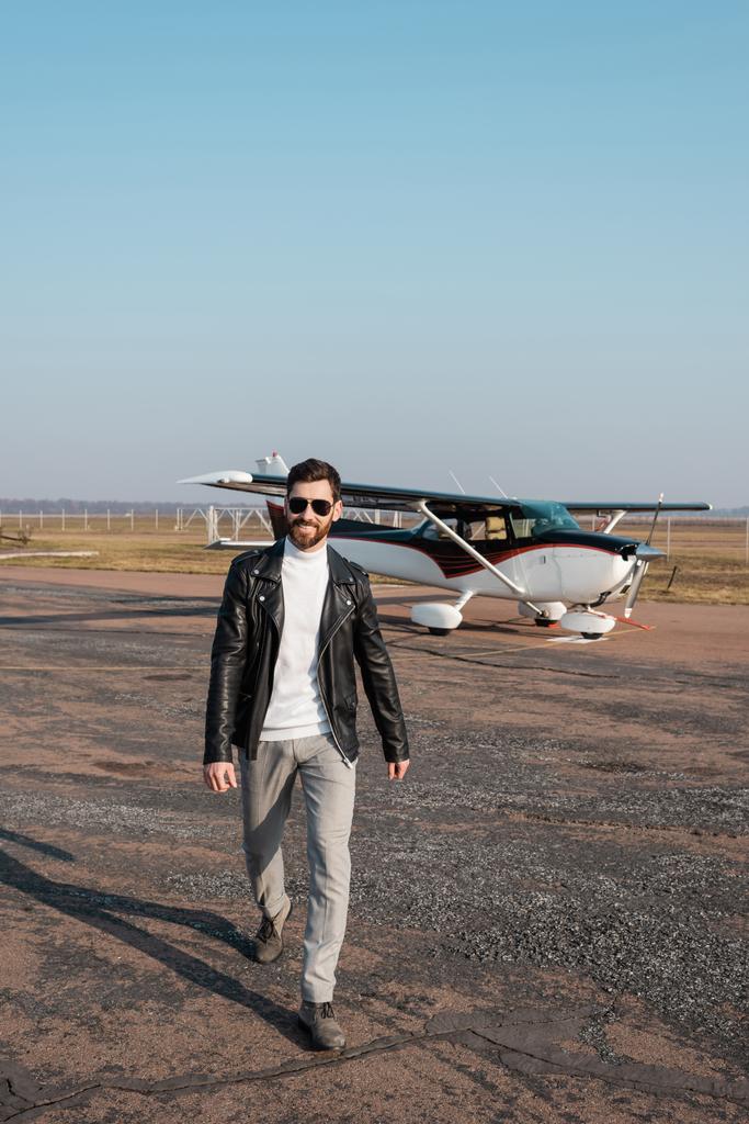 Havalı deri ceketli, güneş gözlüklü, helikopterin yanında yürüyen pozitif pilot. - Fotoğraf, Görsel