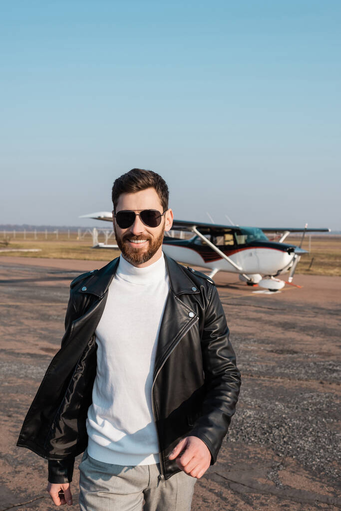 positive pilot in stylish leather jacket and sunglasses walking near helicopter - Photo, Image
