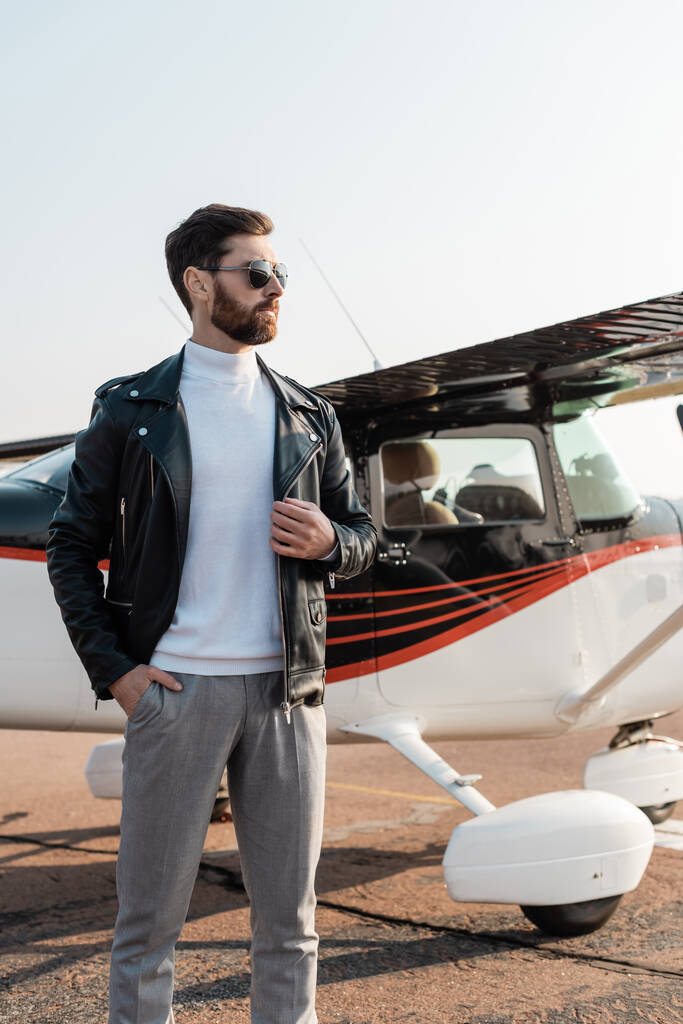 пилот в стильных солнцезащитных очках и кожаной куртке позирует с рукой в кармане рядом с самолетом  - Фото, изображение