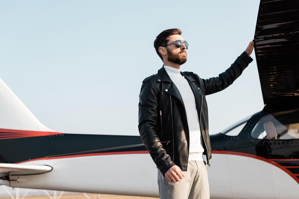 piloto confiante em óculos de sol elegantes e jaqueta de couro em pé perto da asa da aeronave  - Foto, Imagem