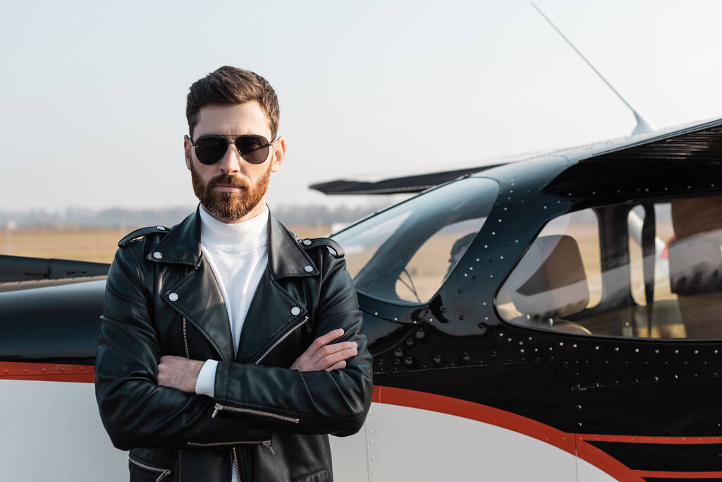 piloto barbudo em óculos de sol elegantes e jaqueta de couro de pé com braços cruzados perto de aeronaves  - Foto, Imagem