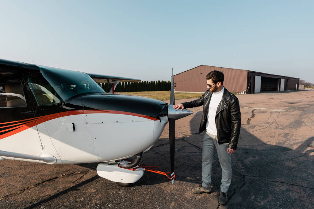 teljes hossza szakállas pilóta stílusos napszemüveg és bőrkabát érintő modern repülőgépek  - Fotó, kép