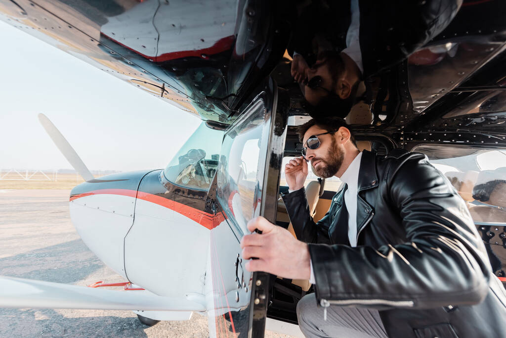 γενειοφόρος πιλότος ρυθμίζοντας κομψά γυαλιά ηλίου και ανοίγοντας την πόρτα στο ελικόπτερο  - Φωτογραφία, εικόνα