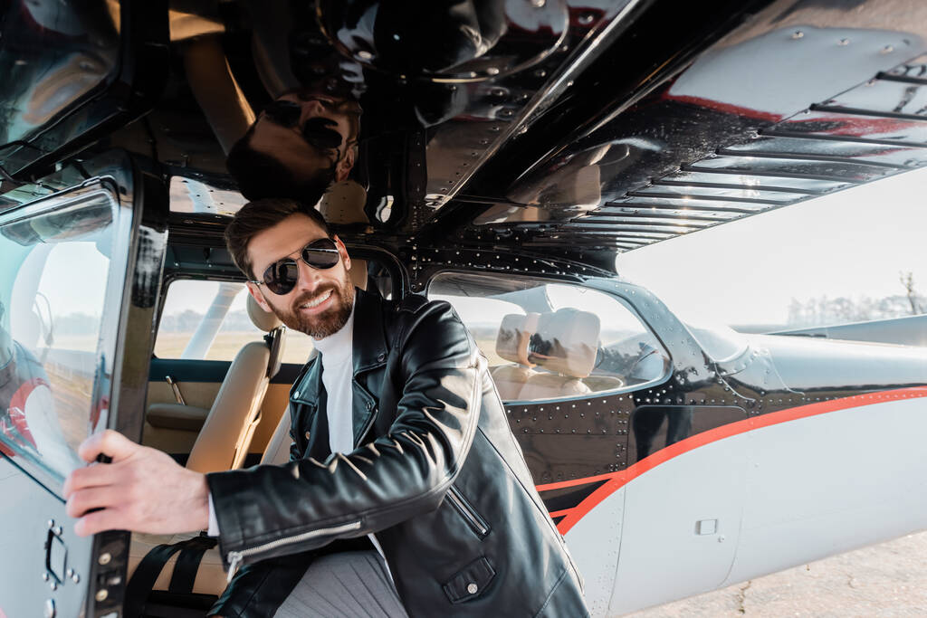 vrolijke piloot in stijlvolle zonnebril en leren jas openingsdeur in helikopter  - Foto, afbeelding