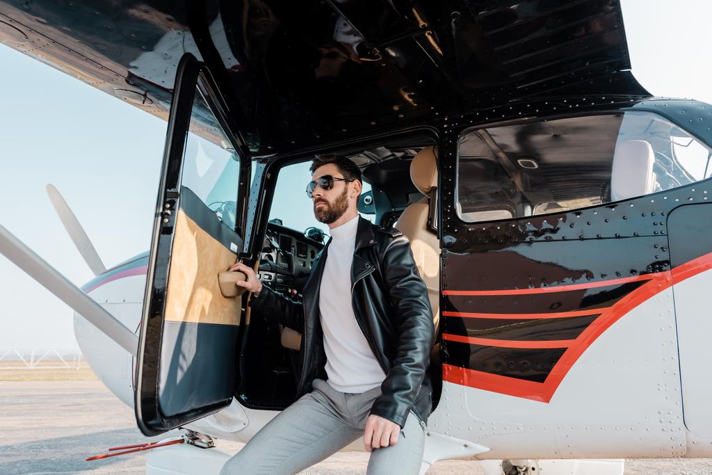 bärtiger Pilot mit stylischer Sonnenbrille und Lederjacke an Hubschrauber gelehnt  - Foto, Bild