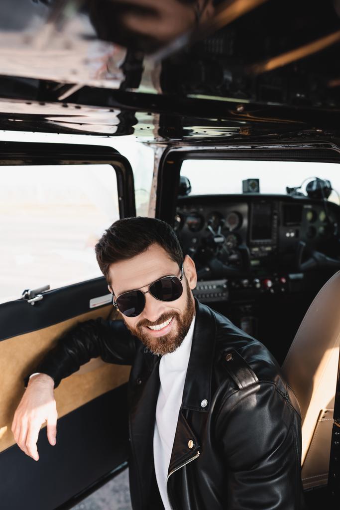положительный пилот в стильных солнцезащитных очках и кожаной куртке возле открытой двери вертолета  - Фото, изображение
