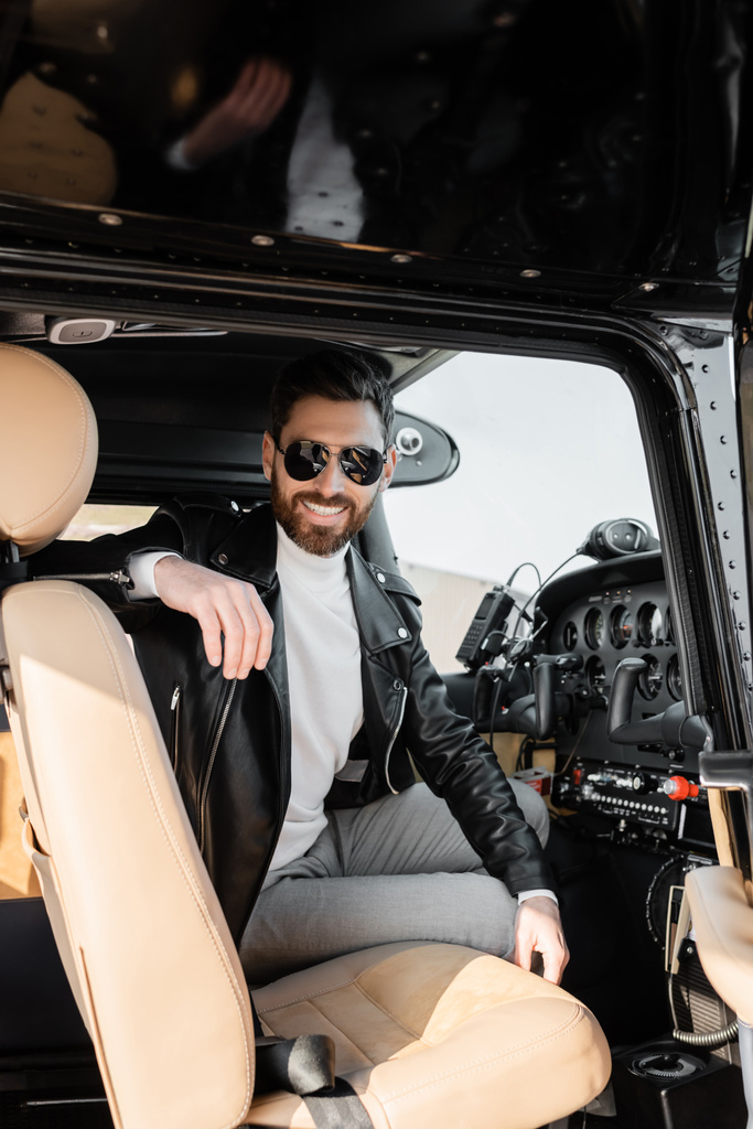 vrolijke piloot in stijlvolle zonnebril en leren jas zittend in cockpit van helikopter  - Foto, afbeelding