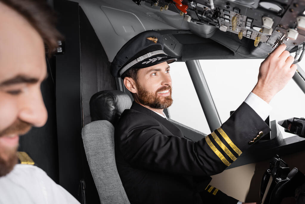 pilota sorridente in uniforme che raggiunge il pannello aereo vicino al copilota offuscato nel simulatore di aeroplano - Foto, immagini