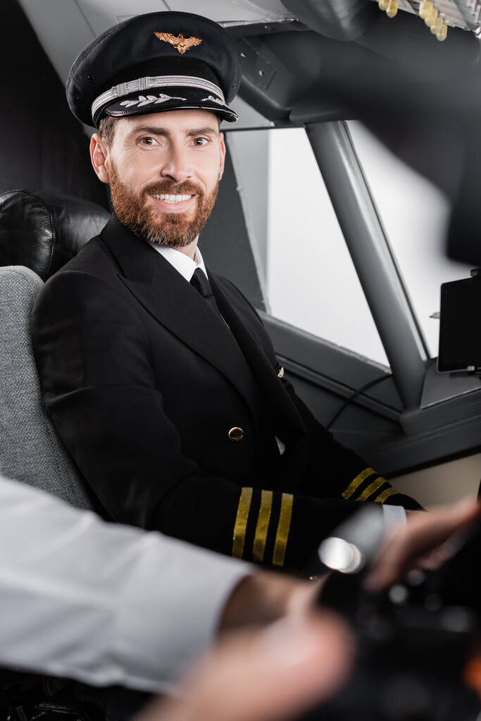 γενειοφόρος πιλότος με στολή και καπέλο χαμογελά κοντά θολή συγκυβερνήτης  - Φωτογραφία, εικόνα