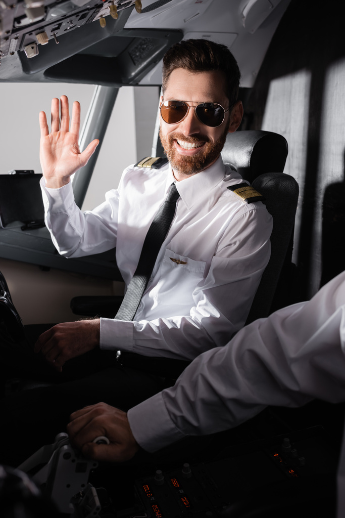 Fröhlicher Pilot mit Sonnenbrille winkt in der Nähe des Co-Piloten im Flugzeug  - Foto, Bild