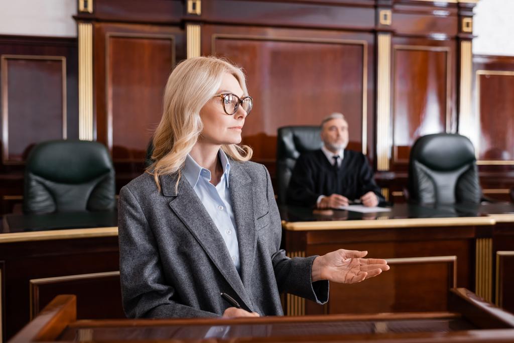  blondynka adwokat wskazując z ręki podczas rozmowy w sądzie w pobliżu sędziego na tle - Zdjęcie, obraz