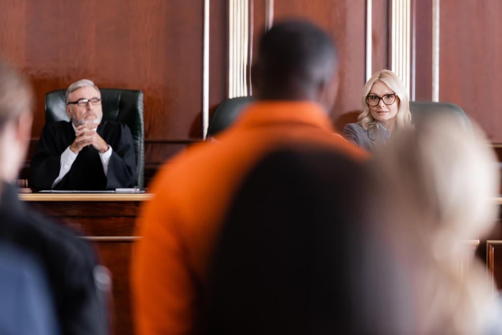 Kıdemli yargıç elleri sımsıkı oturarak avukatın yanında oturuyor ve Afro-Amerikan bir adamı bulanık ön planda suçladı. - Fotoğraf, Görsel
