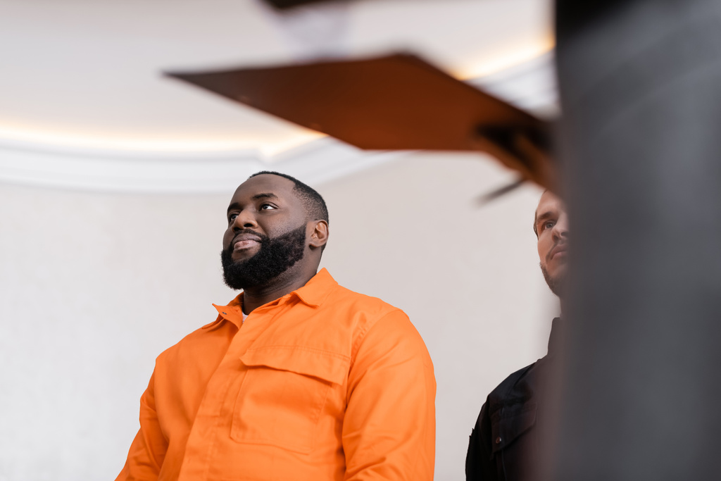 vista de ángulo bajo del hombre afroamericano positivo en uniforme de la cárcel cerca de guardia en la corte en primer plano borroso - Foto, imagen