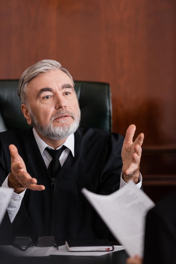 starší soudce ukazuje rukama, zatímco mluví s rozmazaným žalobcem s žalobou - Fotografie, Obrázek