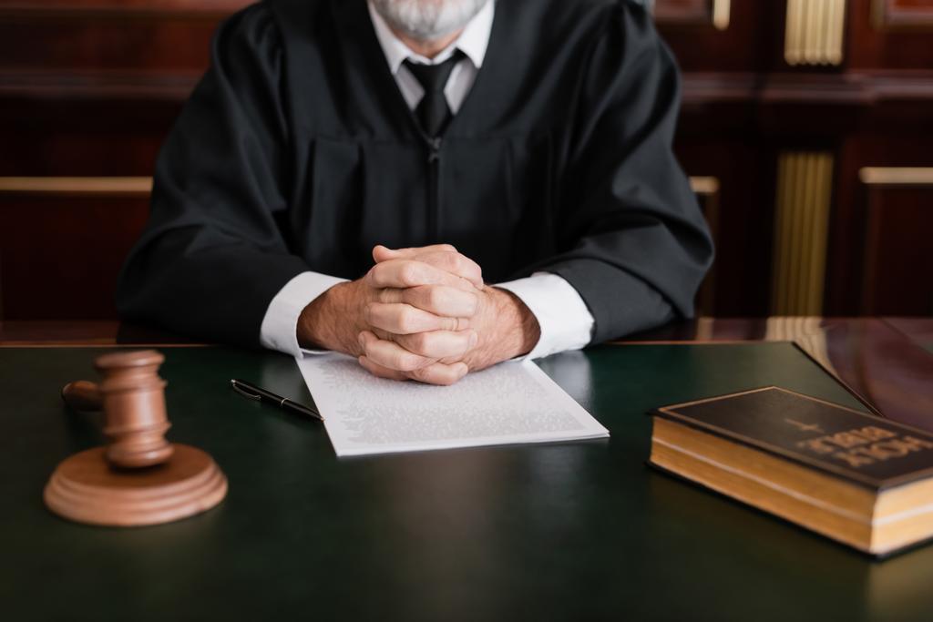 частичный взгляд судьи, сидящего с сжатыми руками рядом с библией, молотком и иском на столе - Фото, изображение