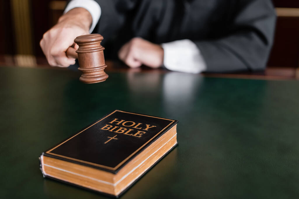 részleges kilátás elmosódott bíró kezében kalapács közelében szent biblia az asztalon - Fotó, kép