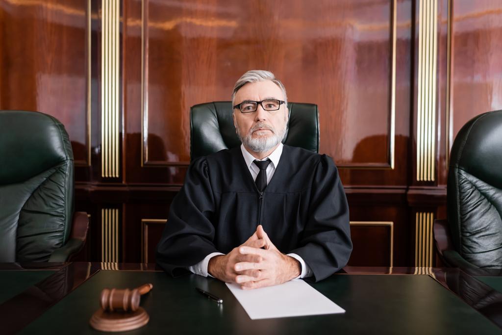 juiz sênior em manto e óculos sentados com as mãos apertadas perto de martelo e papel na mesa - Foto, Imagem